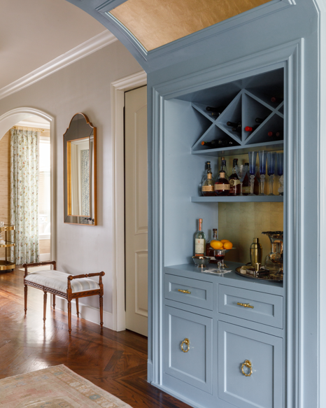 A built-in blue cabinetry and bar station with shelves, wine bottles, and small fixtures, shown in a styled interior room with wood floors and an arched doorway.