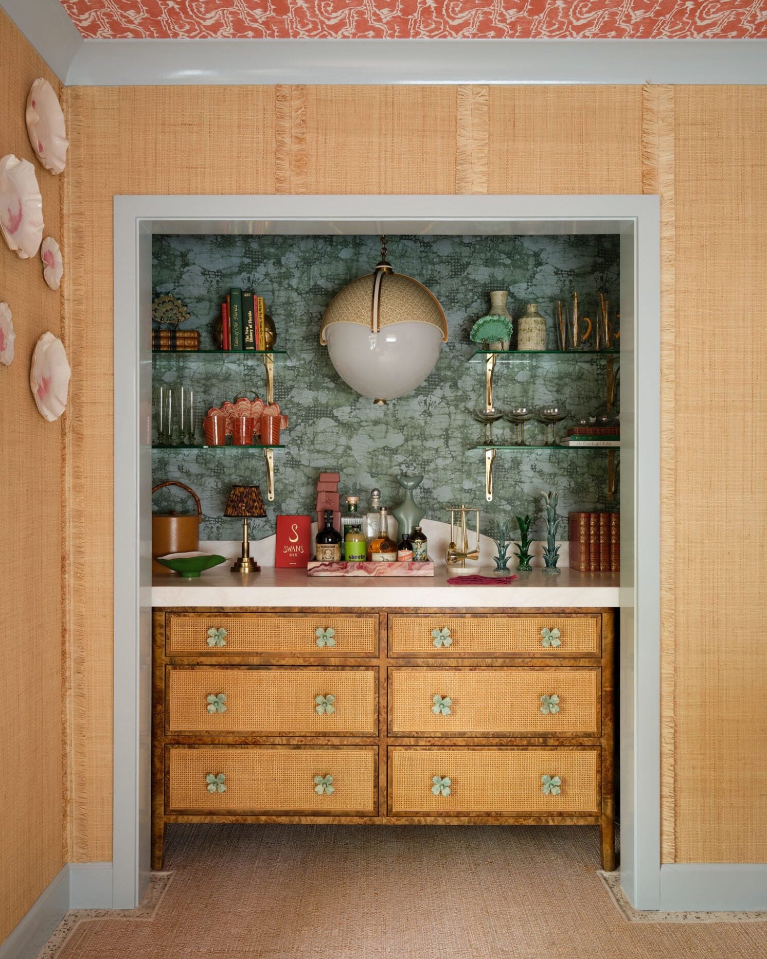 A vintage-style wooden chest of drawers with six drawers and decorative metal knobs, set against a green patterned wallpapered wall in a decorated room.