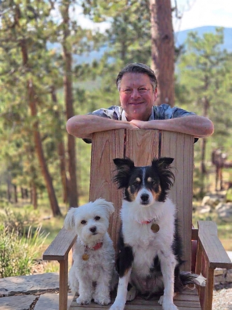 A man smiling and leaning on a wooden chair outdoors with two dogs sitting in front of him. The setting is a wooded area with trees and hills in the background.