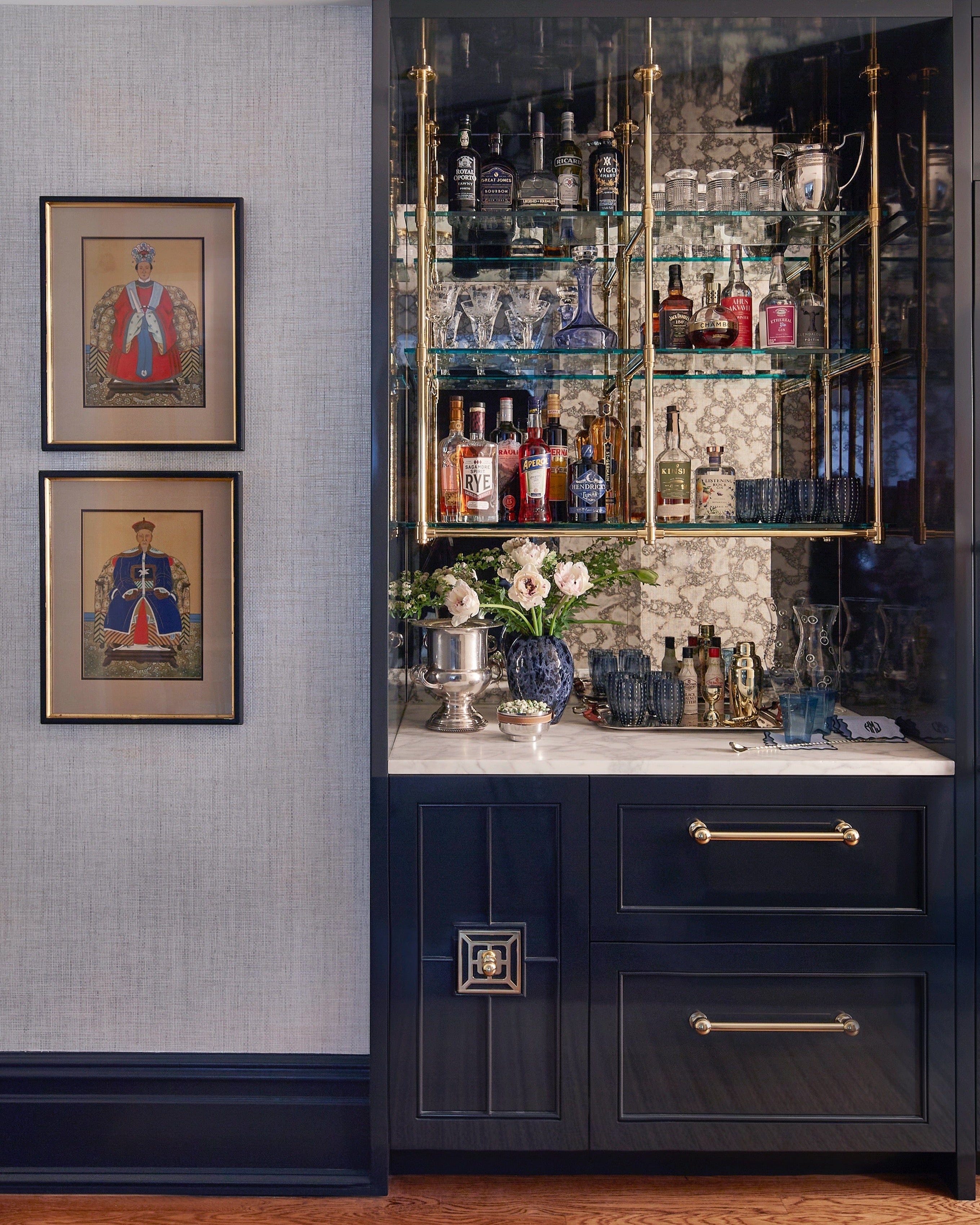 Modern bar setup with shelves of alcohol bottles, glasses, and decorative items against a gray wall with framed pictures.